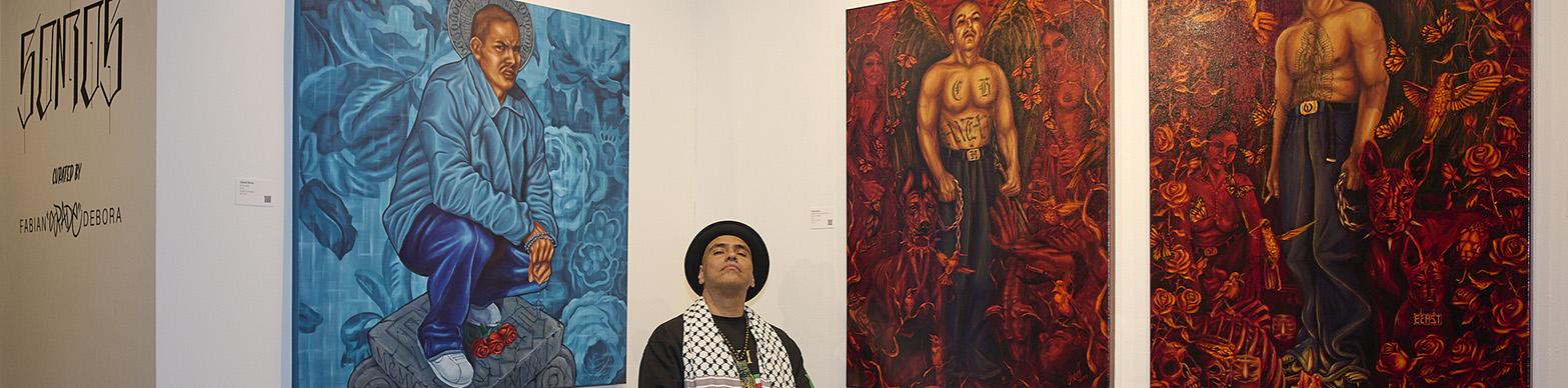 Artist in a wheelchair poses infront of artworks depicting shirtless mexican men on red backgrounds