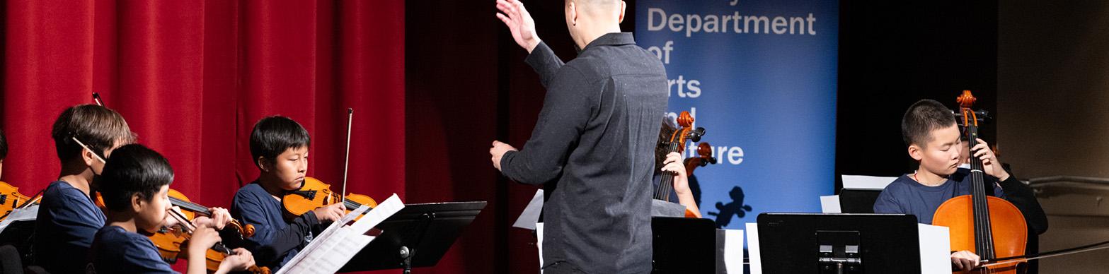 A conductor dressed in all black stands at the center of the stage, facing a row of elementary students dressed in navy T-shirts and dark pants. The students are performing on violin, viola, cello and double bass while referencing sheet music from the stands in front of them.