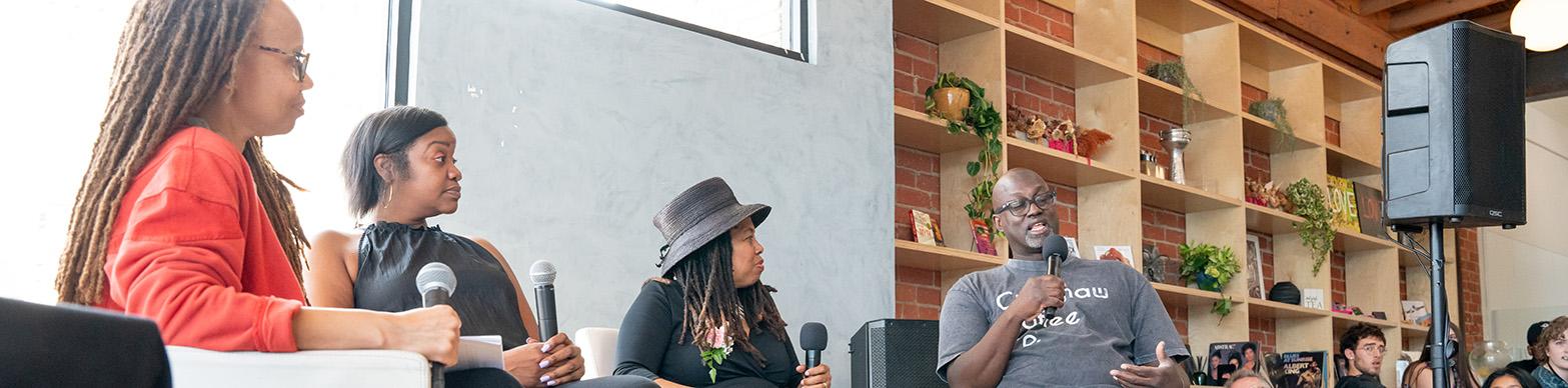 Four individuals sit on stage and chat in front of a live audience.