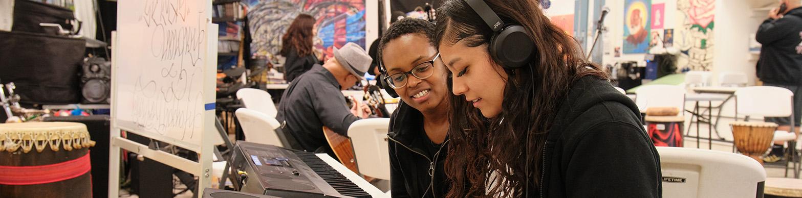 A latina woman plays the piano while a black woman looks on and smiles
