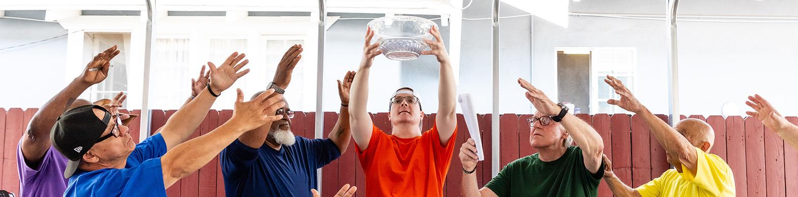 Six older men reach towards a glass bowl held above their heads