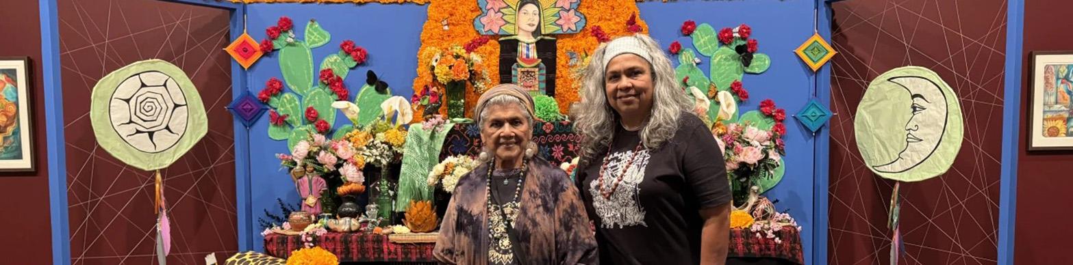 Two older women stand in front of an ofrenda