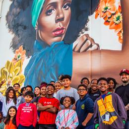 group of children and adults standing in front of a Mural of a black woman dressed as rosie the riveter