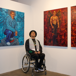 Artist in a wheelchair poses infront of artworks depicting shirtless mexican men on red backgrounds