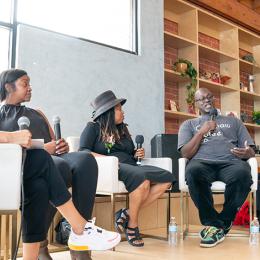 Four individuals sit on stage and chat in front of a live audience.